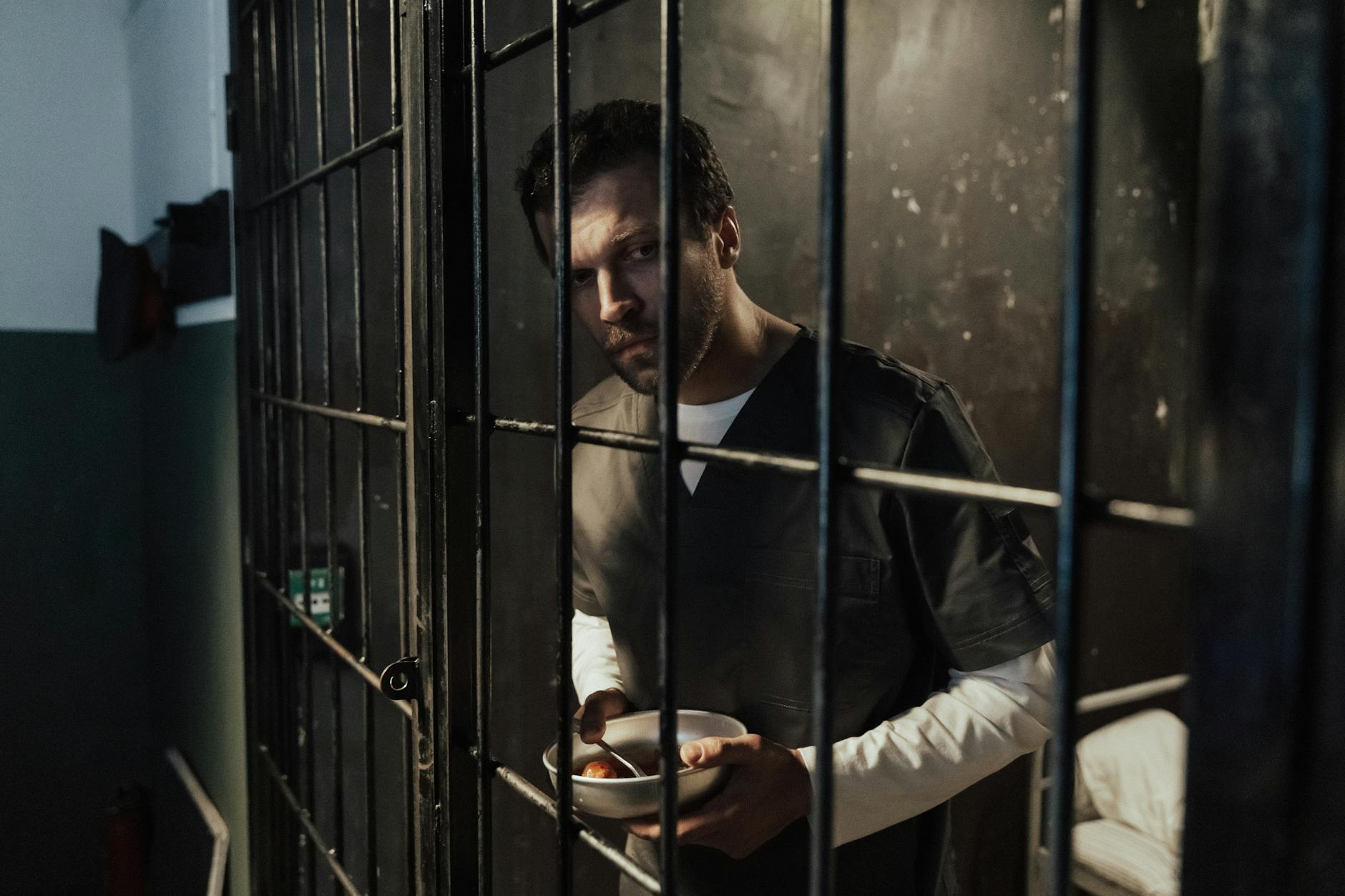 Adult male prisoner holding a meal inside a dimly lit jail cell, looking thoughtful.